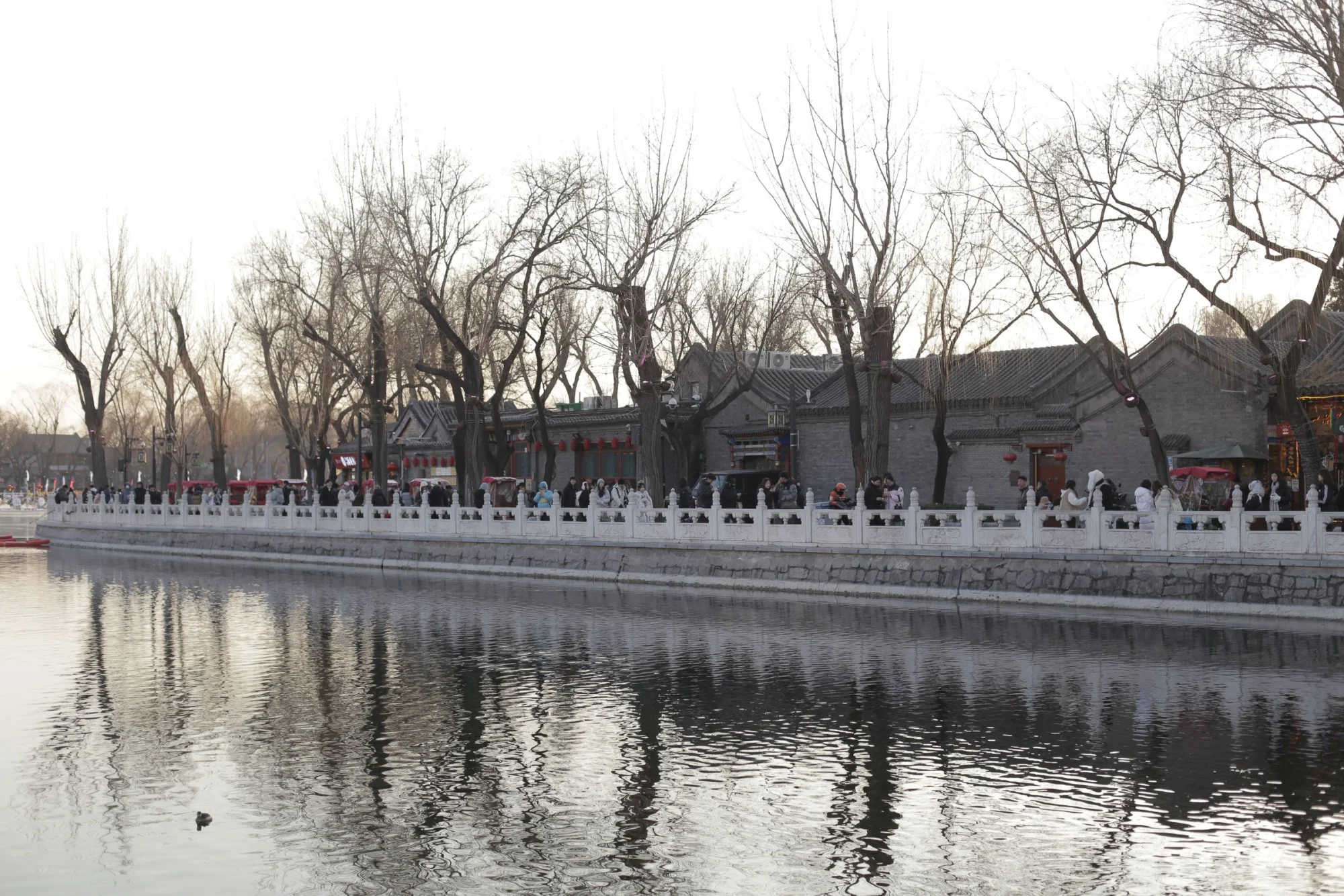 Daily life along Shichahai lakeside in Beijing with bare winter trees and traditional architecture reflecting on the water.