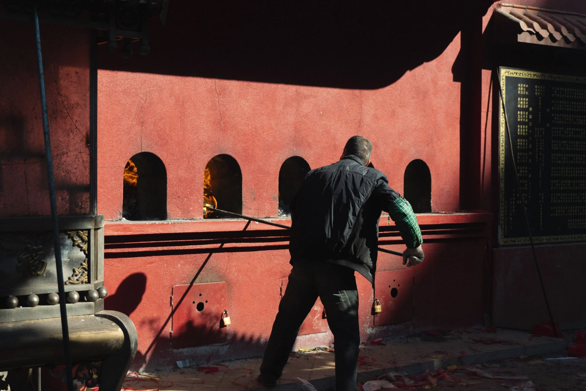 A man lights incense at an outdoor temple shrine, casting shadows on a red wall.