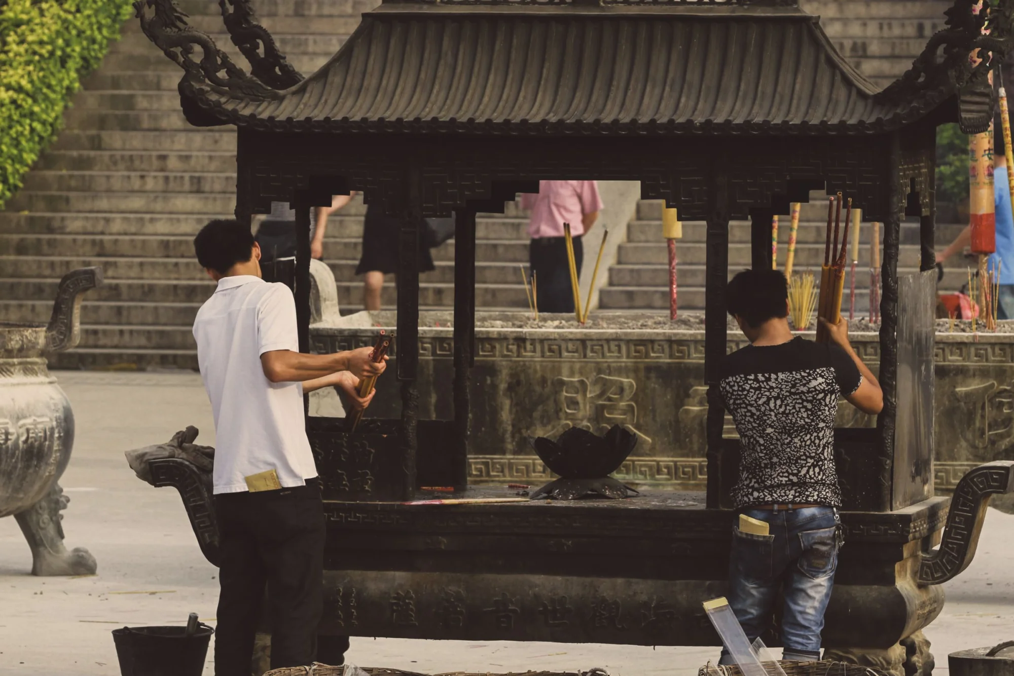 Two men light incense sticks at an outdoor shrine, symbolizing tradition and spirituality.