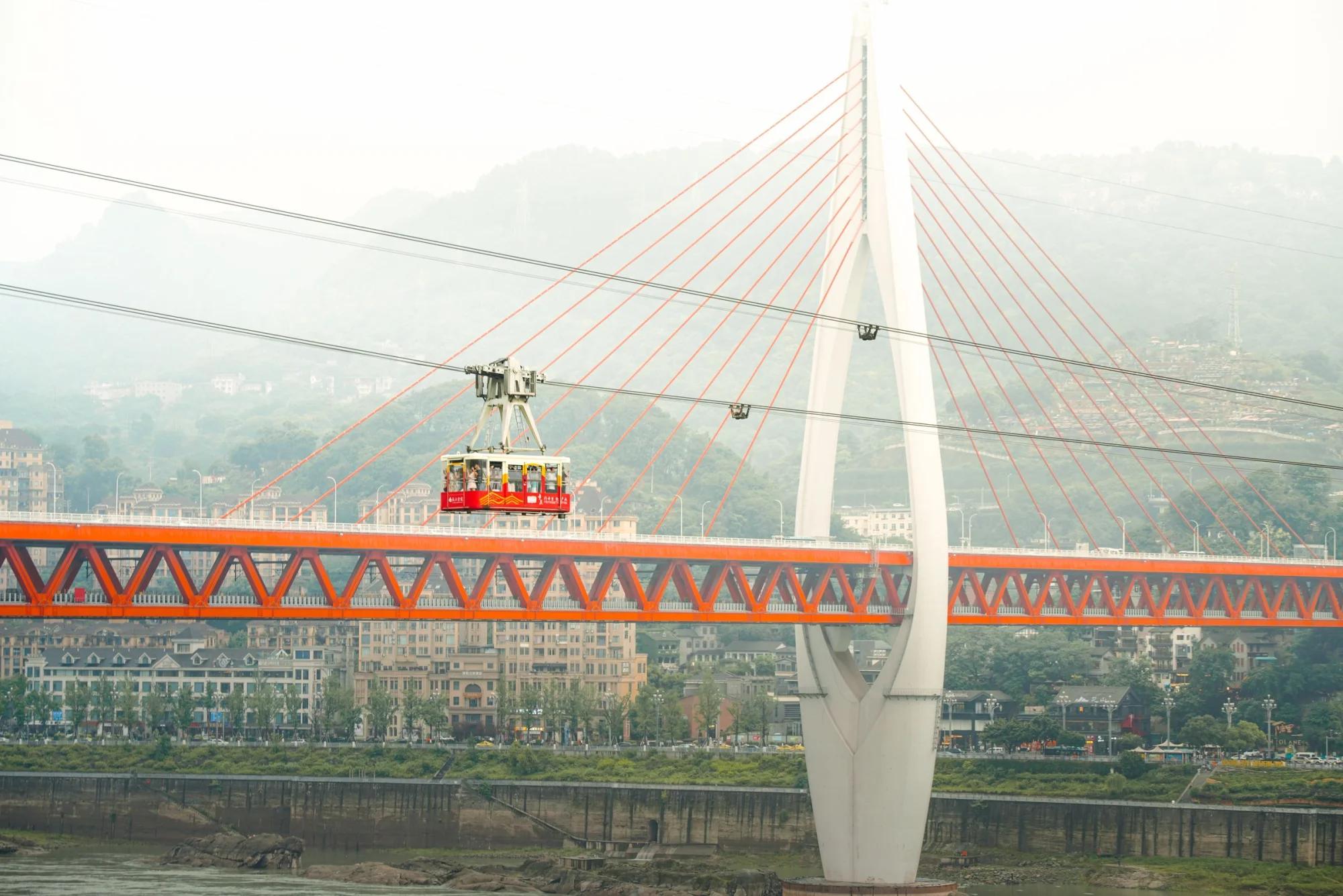 A cable car crosses the Yangtze River bridge in Chongqing, featuring vibrant architecture and lush hills in the background.