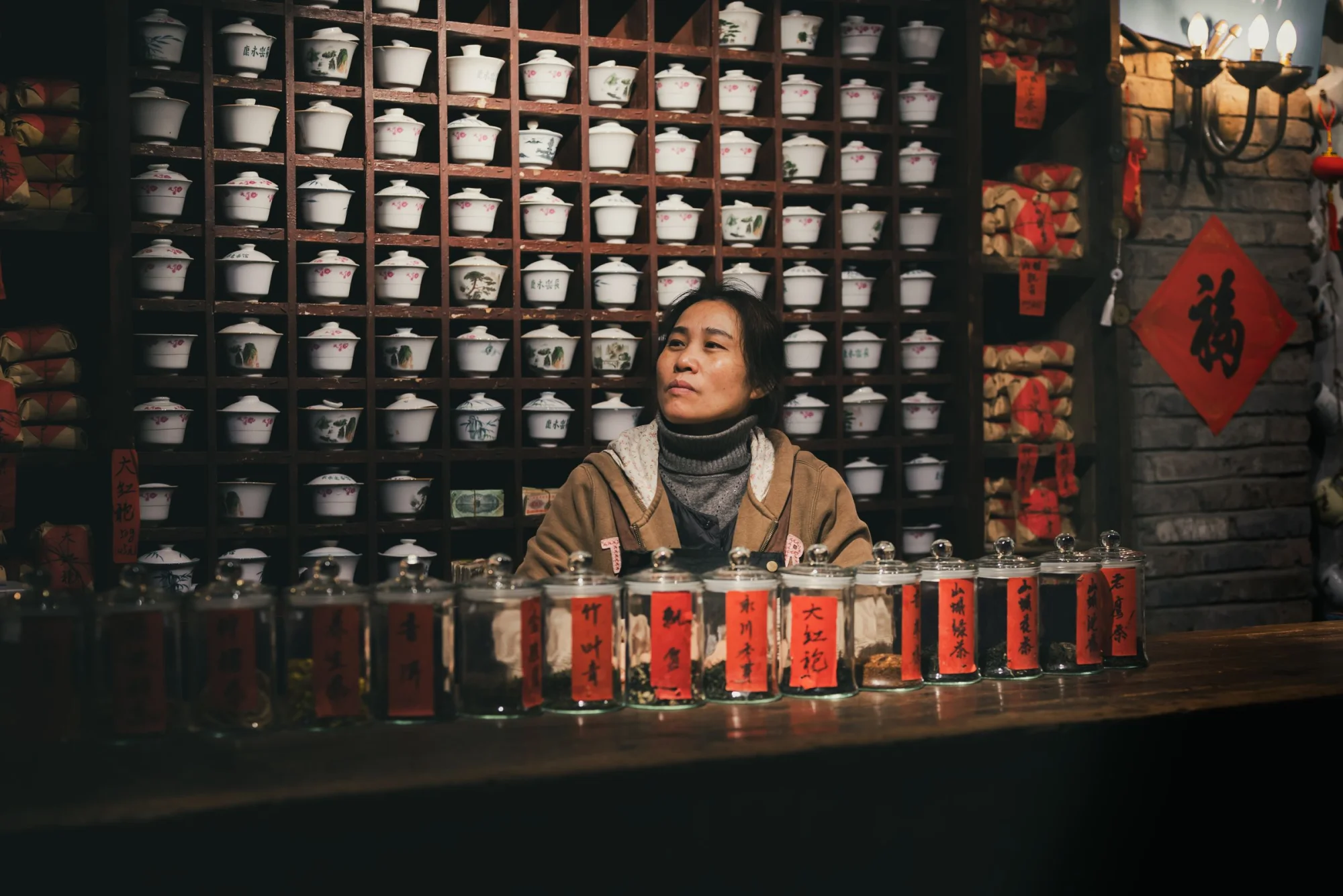 An attentive woman waits in a traditional Chinese teahouse surrounded by jars of tea and teacups on shelves.