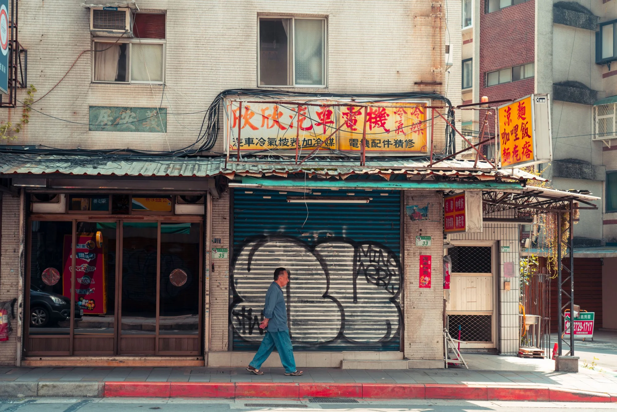 A lone pedestrian walks past a graffiti-covered storefront in an urban setting during the day.