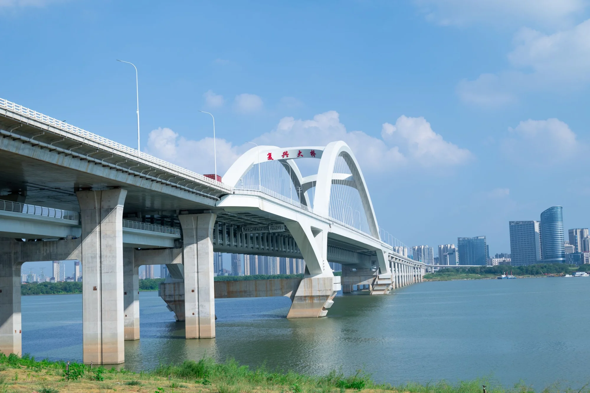 A modern arch bridge spanning a river against a city skyline with clear blue skies.