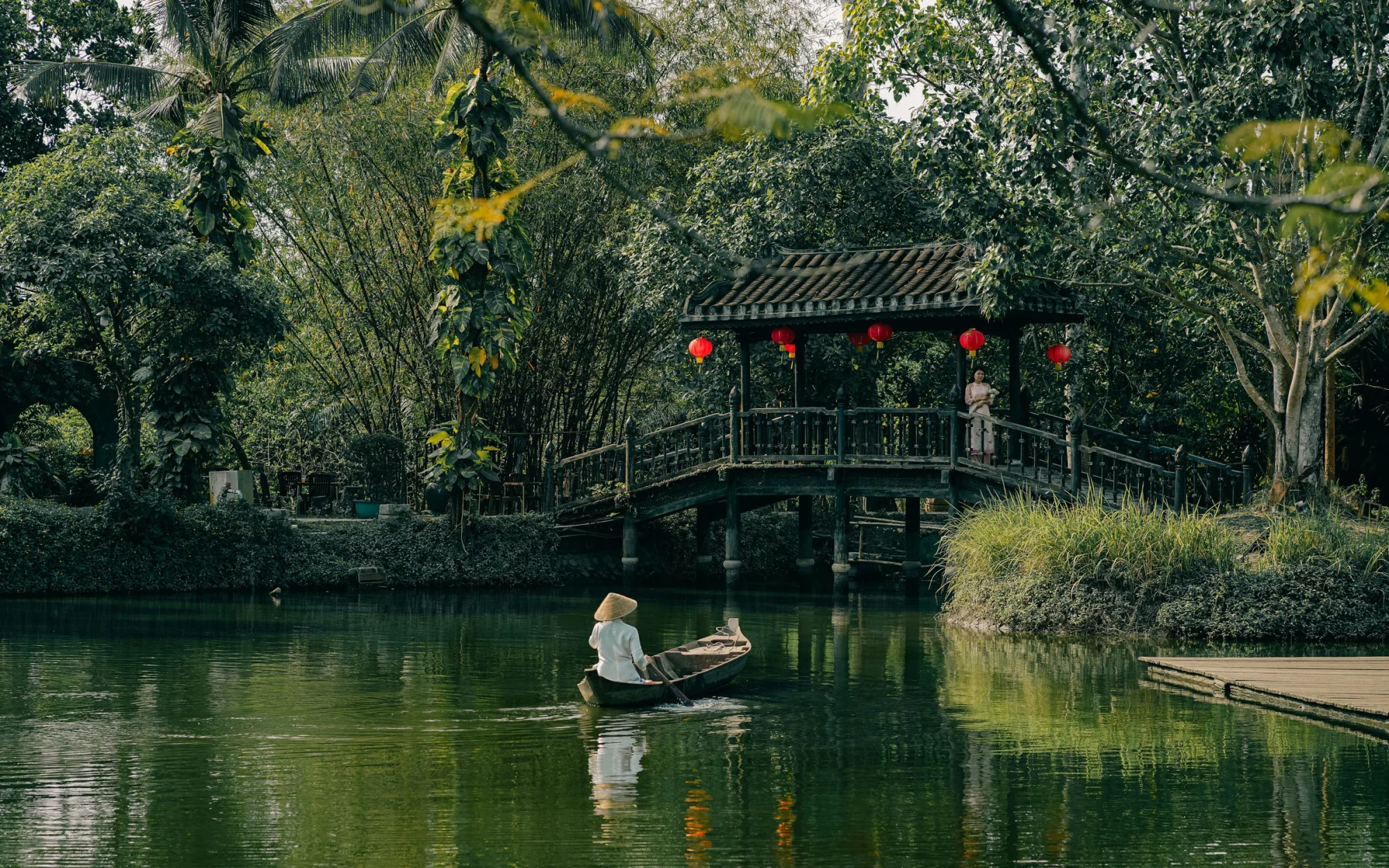 A serene image of a person boating on a pond with a charming footbridge in the background.