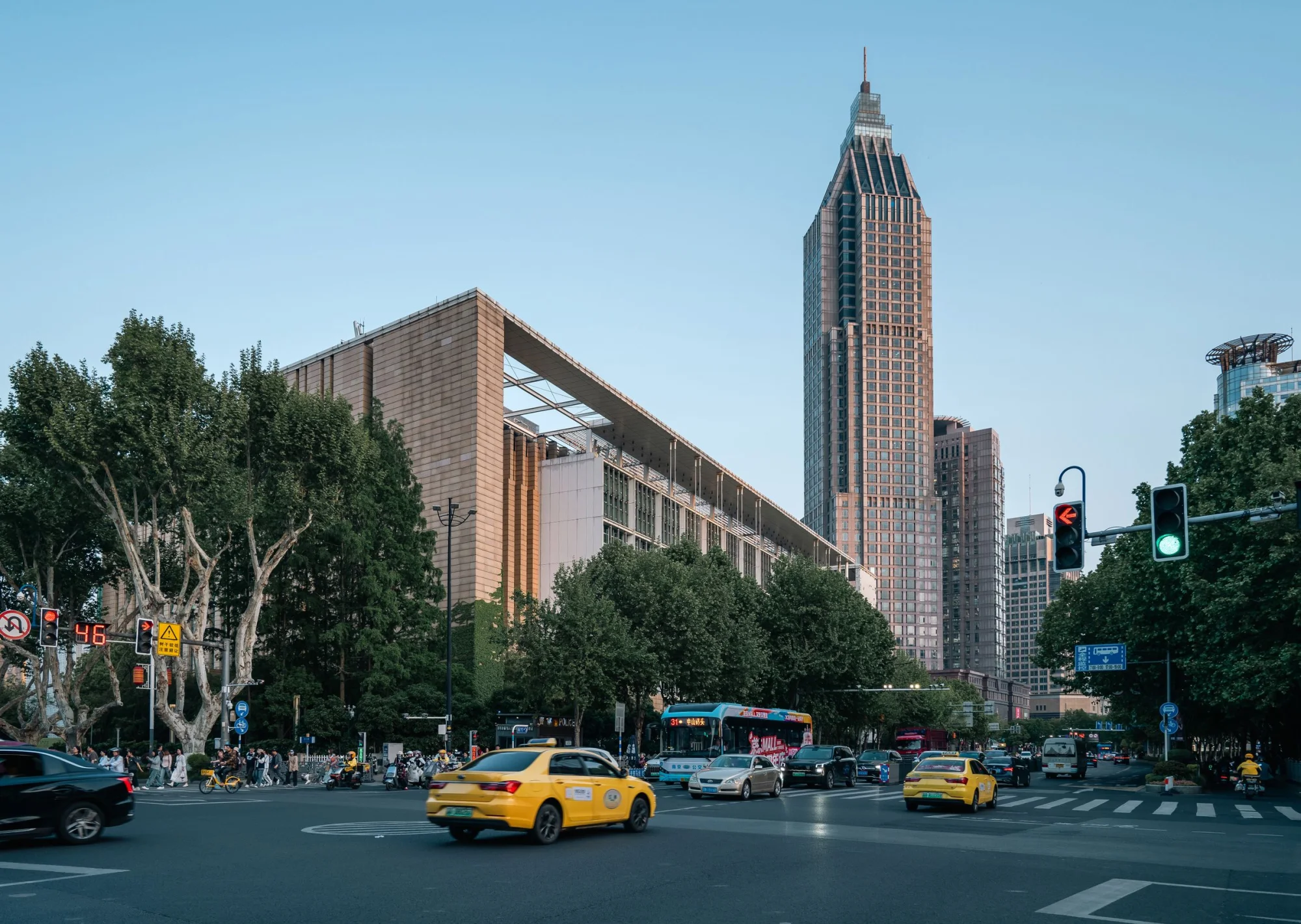 A bustling street scene near Nanjing Library with taxis and a towering skyscraper under a clear sky.