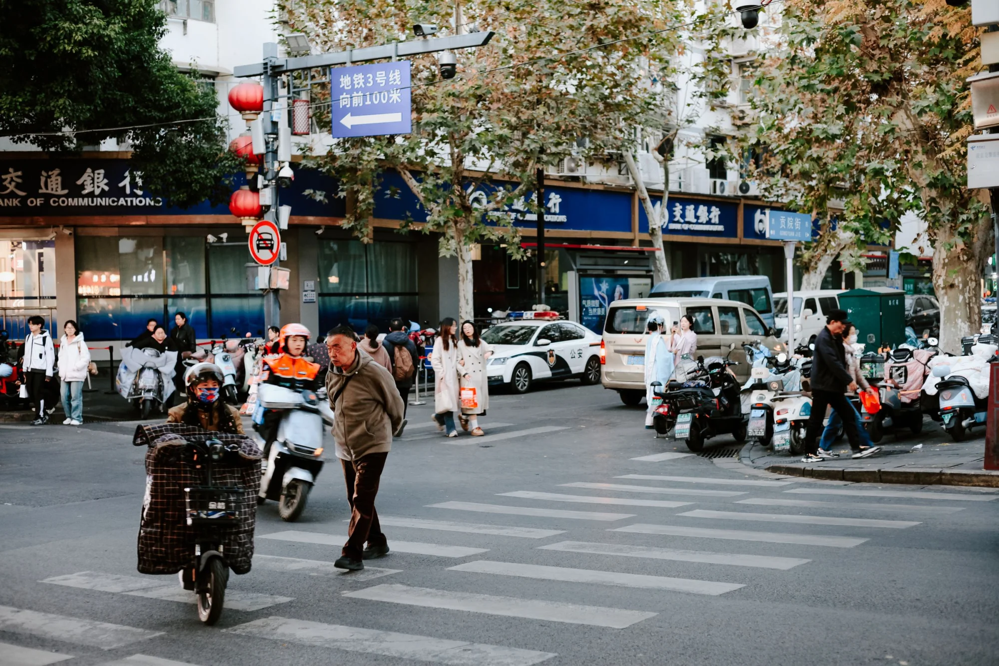 Street life in Nanjing, China with people, scooters, and autumn foliage on a busy corner.