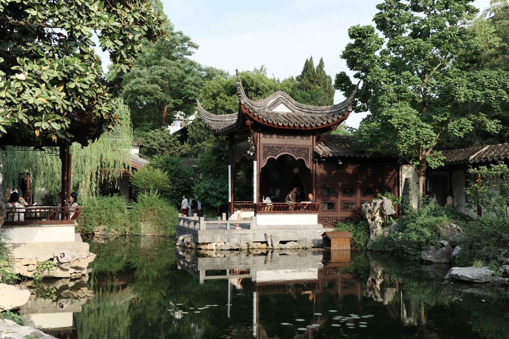 A traditional Chinese pavilion reflected in a tranquil garden pond surrounded by lush greenery.
