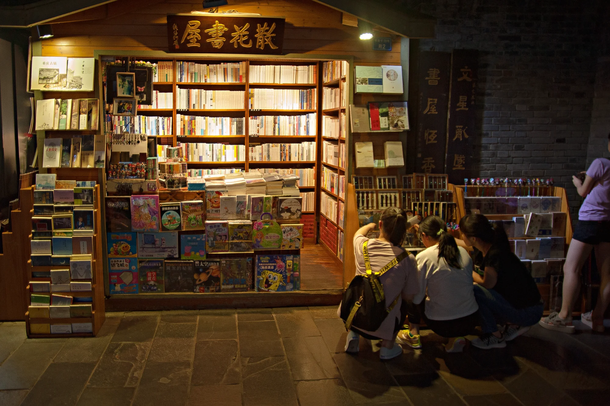 Cozy street bookstore at night with people browsing books and gifts.